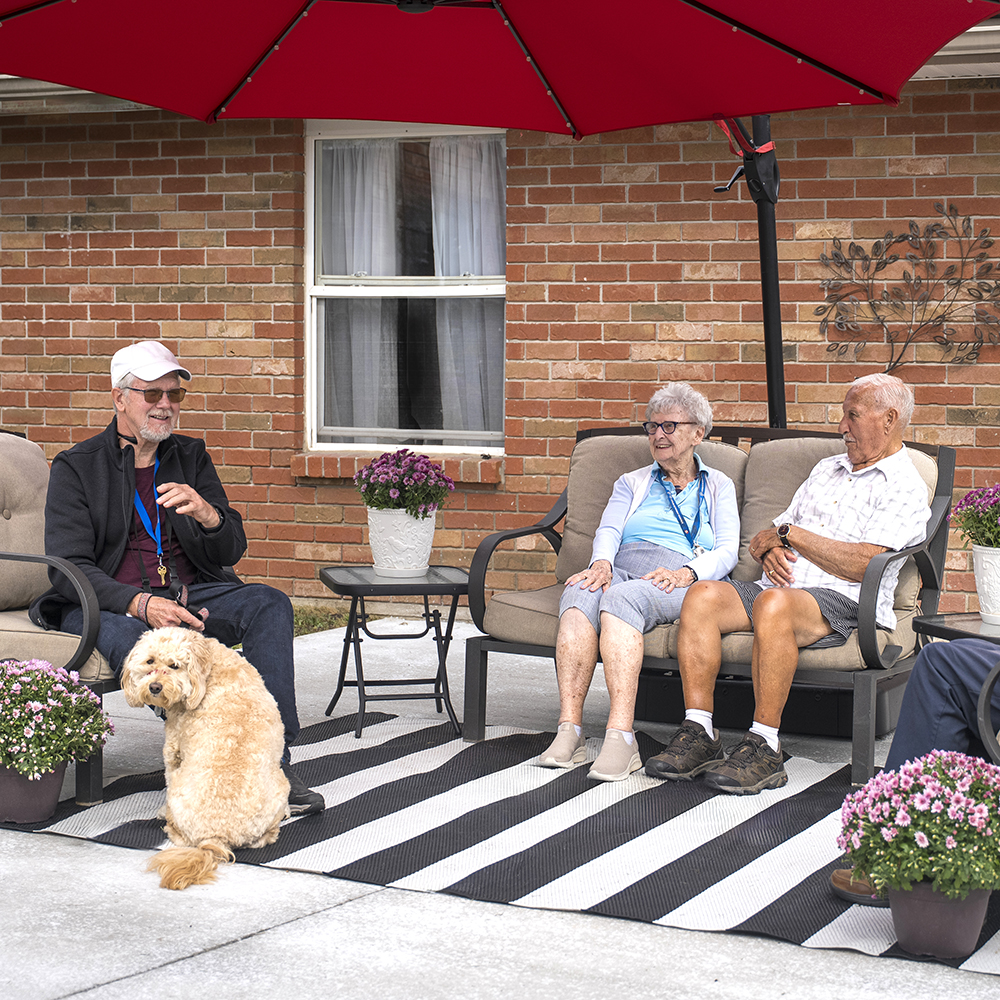 Three residents relaxing outdoors on the patio with a friendly dog on a sunny afternoon at Atrium Retirement Residence in Orillia.